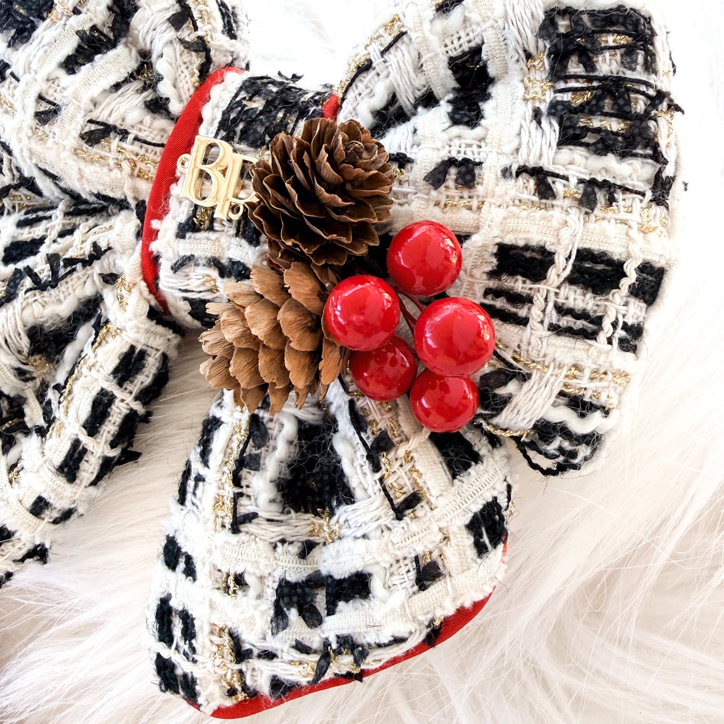 A close-up of a decorative dog bow made from black and white tweed fabric with red trim, adorned with pinecones and red berries, resting on a white faux fur surface.