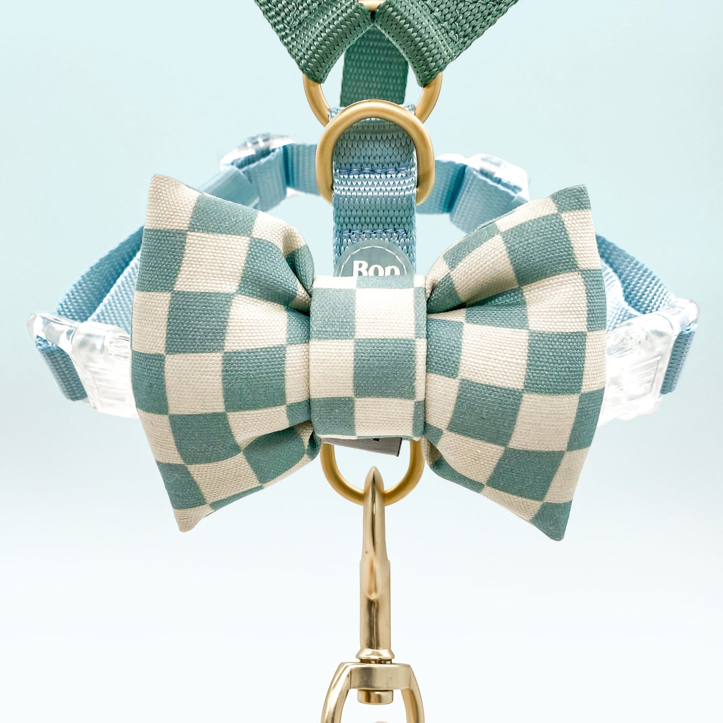 A close-up of a pet harness with a matching checkered bow tie, attached the top park of harness and leash set. The harness and leash are made of a teal and sage green poly fabric with gold-tone metal hardware rings and clips.