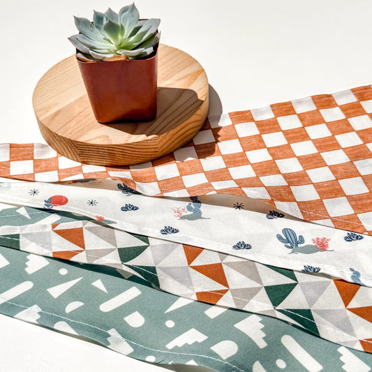 Four pet bandanas patterns arranged diagonally on a white surface. Top bandana features a terracotta and white checkerboard pattern, middle is white with blue, green, red cactus and star print, and bottom bandana is sage green with white abstract shapes.