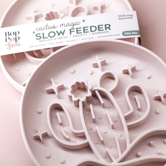 actus Magic Slow Feeder staggered on muted, dusty pink colored background with top feeder displayed with brand packaging labeled 'Cactus Magic SLOW FEEDER' and lower displaying close up details of cactus and stars 3 dimensional pattern in dramatic lighting showing a blush milky beige color.