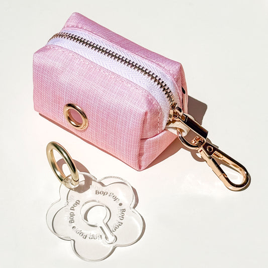 A small pink texture-printed waterproof fabric zipper pouch with a gold-tone metal grommet and attached gold-tone clasp, shown on a white background with a clear flower-shaped poop loop engraved with 'Bop Pop' for dog poop bag dispensing.