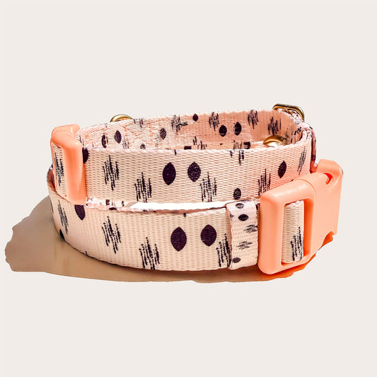 A dog collar with a ikat pattern of black abstract marks on a light peach-pink background, featuring a bright peach-pink plastic buckle and keeper, photographed on a light beige background.