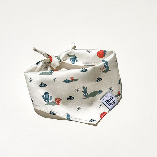 A square dog bandana with a tie closure, featuring a cactus and desert plant pattern on a cream background, with a visible 'Bop Pop' brand label.