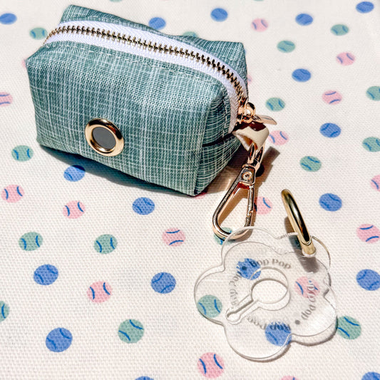 A small zippered pouch in a sage green white textured fabric with a gold-tone metal ring and clasp, shown on a white fabric background patterned with pink, blue, and teal baseballs and balls. A clear flower-shaped tag with gold hardware is placed in front.