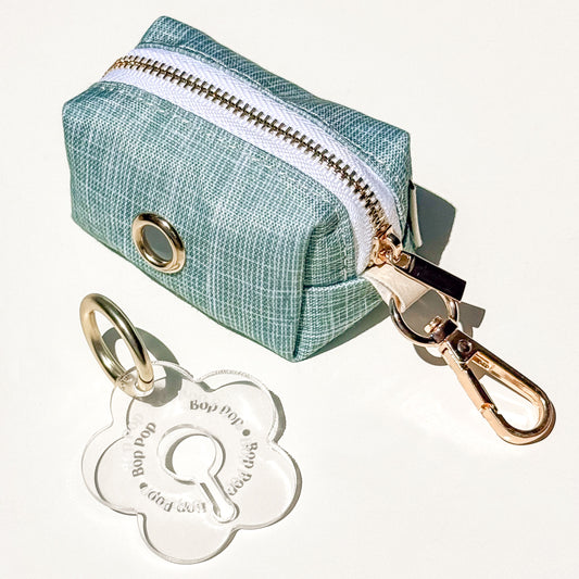 A small sage green texture printed waterproof fabric zipper pouch with a gold-tone metal grommet and attached gold-tone clasp, shown on a white background with a clear flower-shaped poop loop engraved with 'Bop Pop' for dog poop bag dispensing.