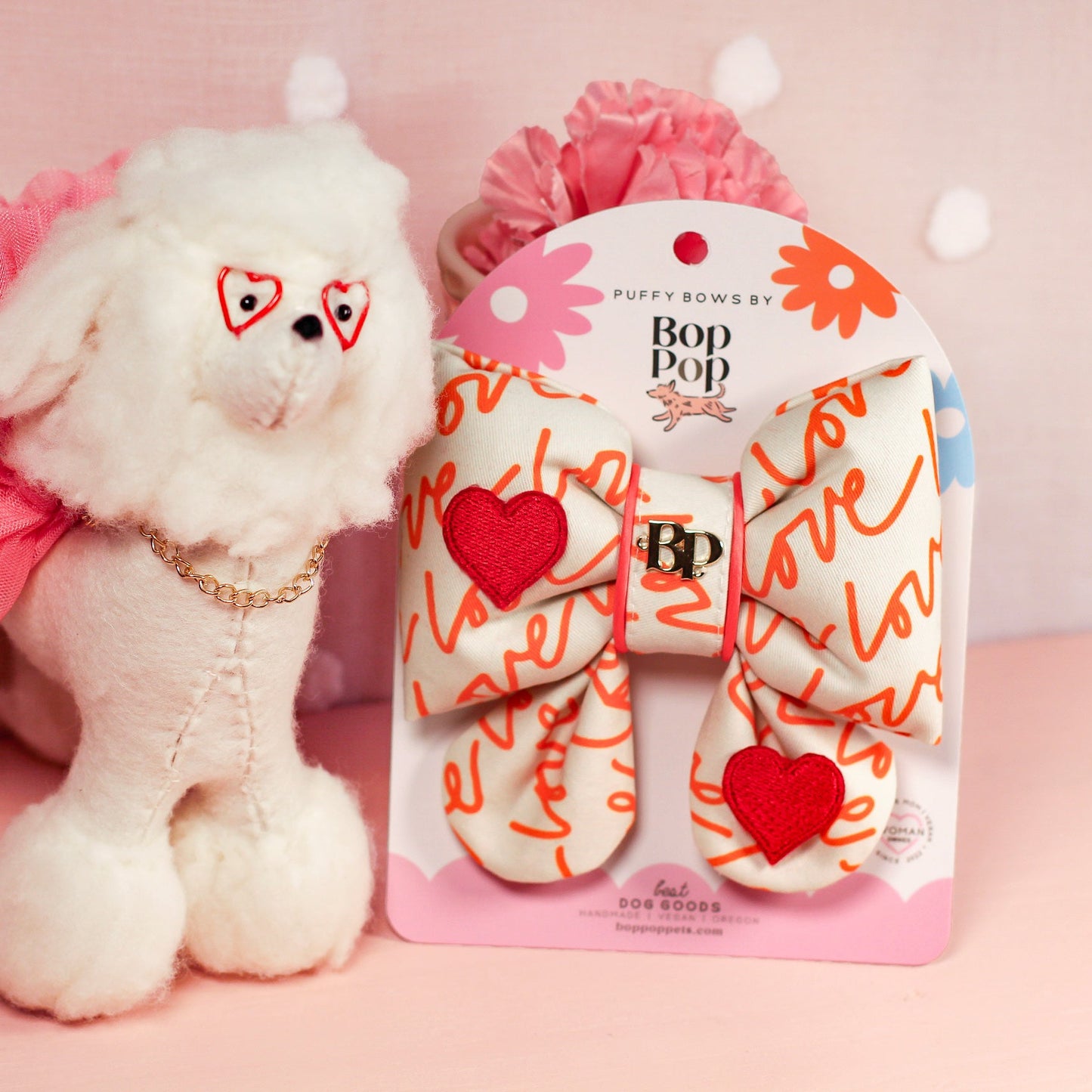 White plush dog toy with pink bow and heart design next to its packaging on a pink background.