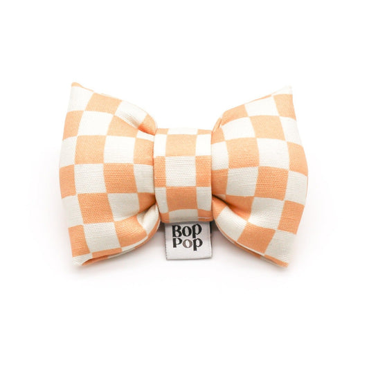 A peach and white checkerboard patterned bow tie for dogs with the brand label 'Bop Pop' visible.