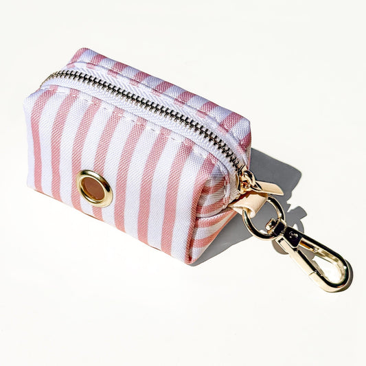 A small zippered pouch with vertical pink and white stripes, featuring a gold-tone metal open grommet and a gold-tone carabiner clip, shown on a clean off-white background.