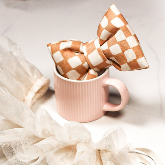 Tan maple checkered bow tie in a pink espresso cup on a marble surface. Bop Pop Pets pet accessory.
