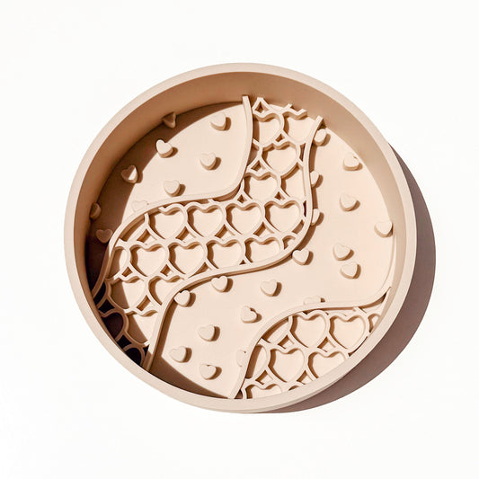 A round beige silicone pet slow feeder bowl with a raised wavy pattern of hearts, photographed from directly above on a clean white background.