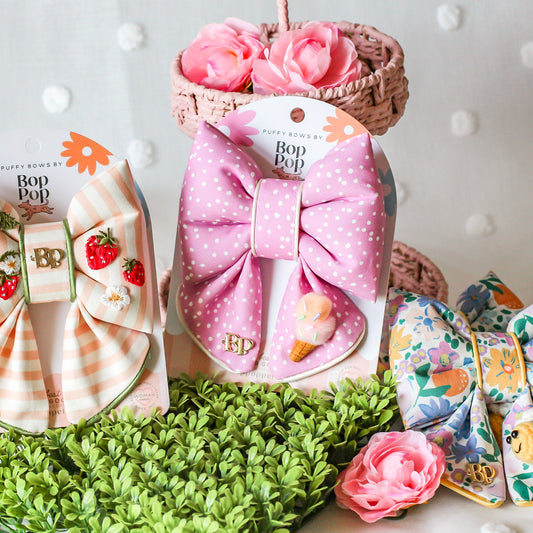 Pink polka dot headband with bow in a box, surrounded by decorative items on a textured surface. Pink polka dot sailor bow tie for dog cats with cute sunglasses, flowers and white satin trim piping. bop pop pets pet accessory