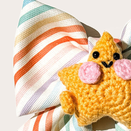 A colorful pastel striped cotton canvas fabric pet bow with a yellow-orange crocheted star character accessory in the center, photographed on a plain white background.