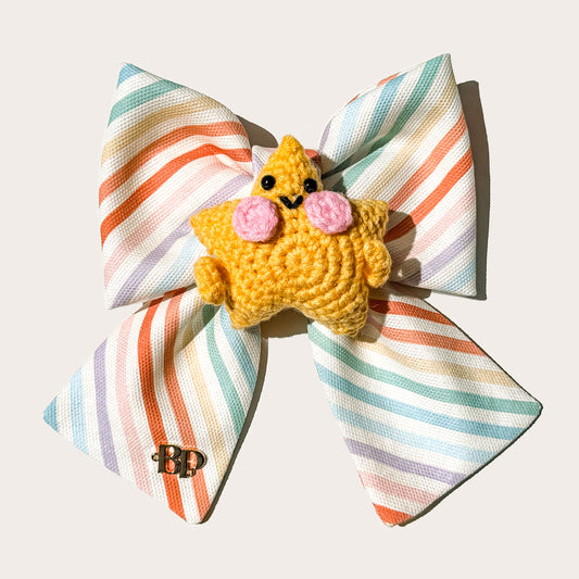 A colorful pastel striped cotton canvas fabric pet bow with a yellow-orange crocheted star character accessory in the center, photographed on a plain white background.