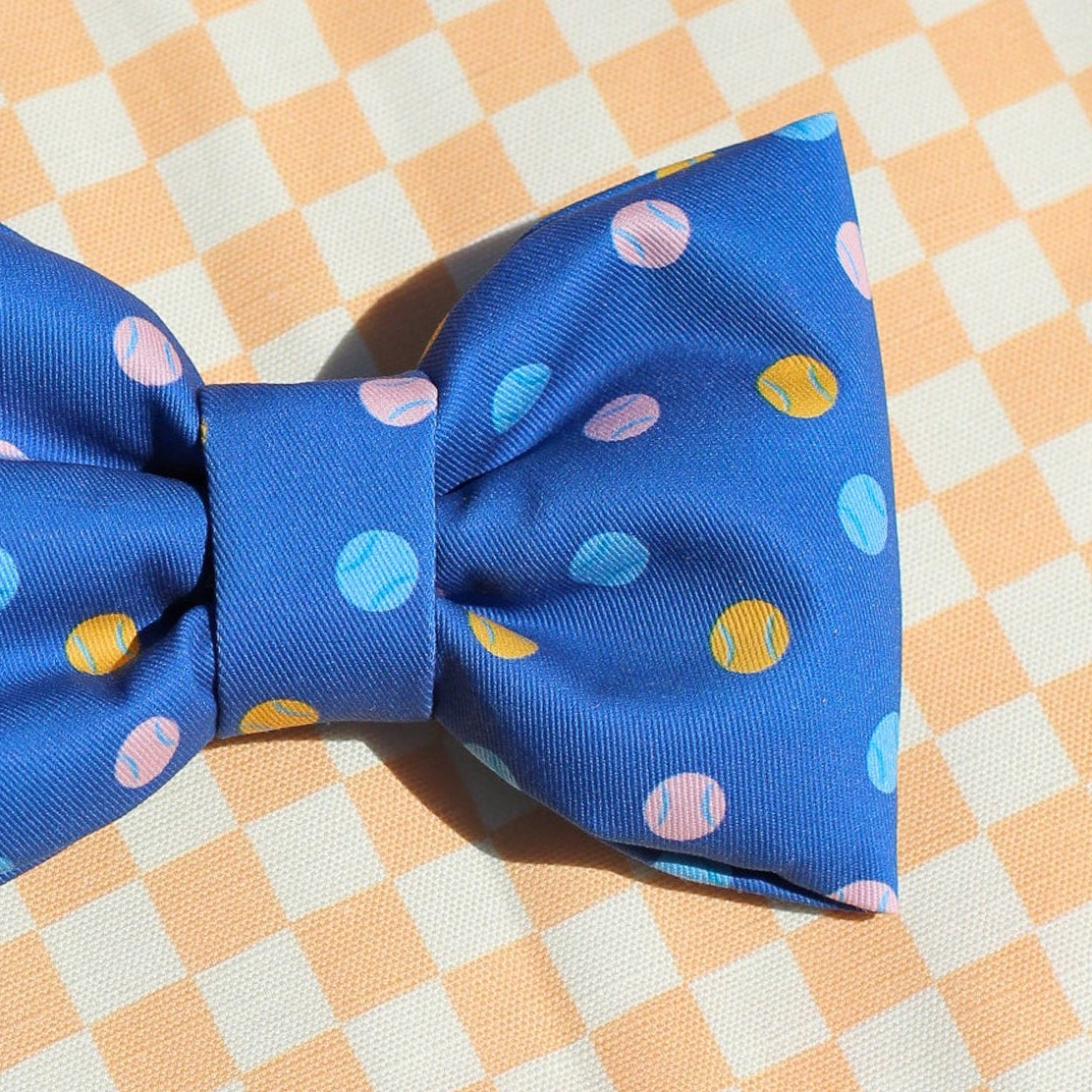 Close up print of Blue bow tie with colorful tennis balls on a dog bow tie on a checkered fabric background for Bop Pop Pets all cotton, puffy bow ties for fashion, trendy dogs cats