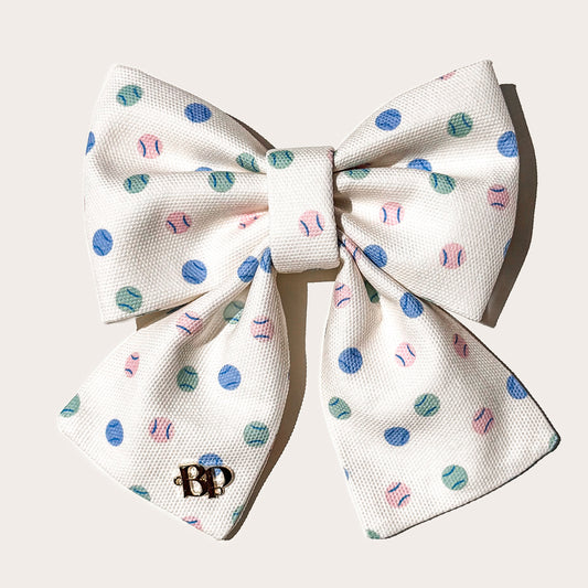 A cotton canvas textured fabric pet sailor bow in cream with a pattern of blue, teal, and pink tennis balls, featuring a small gold 'BP' monogram brand logo on left bow tails on white background.