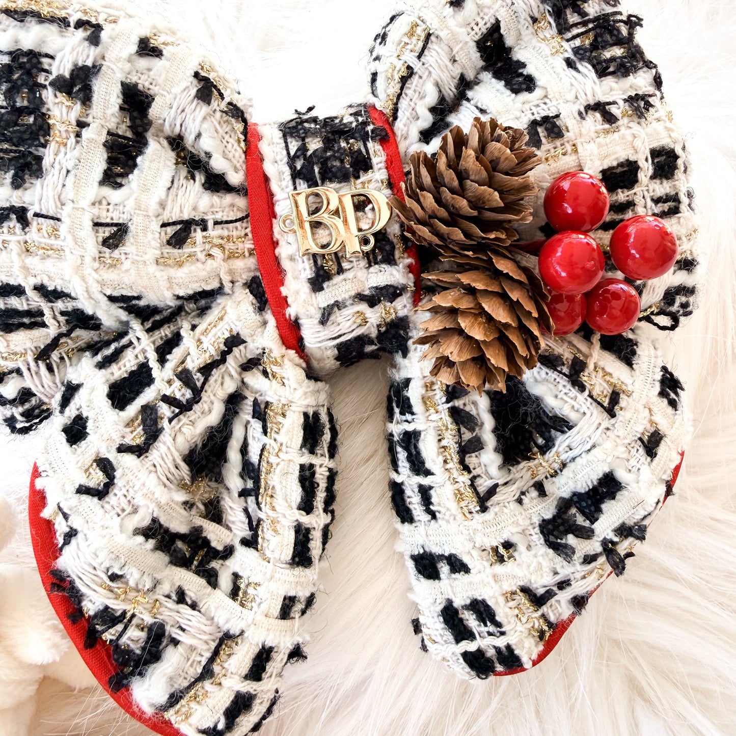 A large decorative pet bow made from black, white, and beige tweed fabric, accented with red trim, a gold brand logo 'BP', a pinecone, and red berries, displayed on a white faux fur background.
