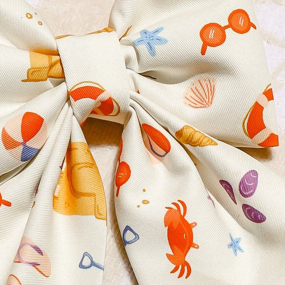 Decorative items including a sun-patterned fabric, crochet sun, and fabric bows on a textured surface. Colorful dog bow with ocean-themed design on a textured surface. Bop Pop Pets for trendy dogs and cats