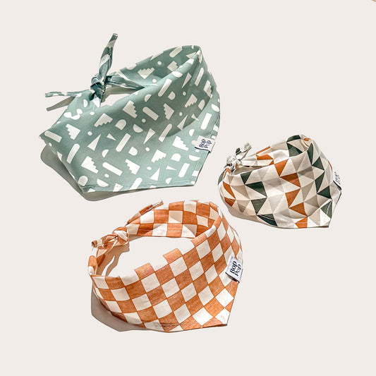 Three patterned pet bandanas arranged on a light beige background. One bandana is sage green with white abstract shapes, one is orange and white checkerboard, and one is cream, green, and orange geometric.