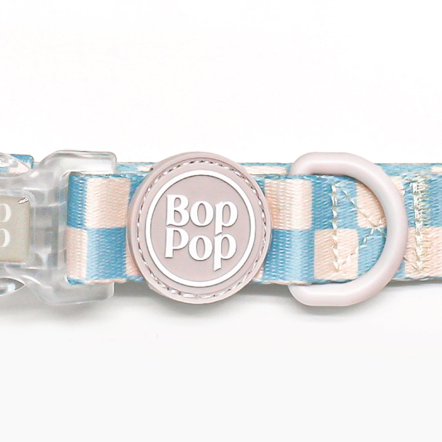 A close-up of a Bop Pop brand dog collar featuring a pastel blue and cream checkered pattern, a light pink plastic D-ring, and a central round pink tag with the brand name 'Bop Pop' in white.