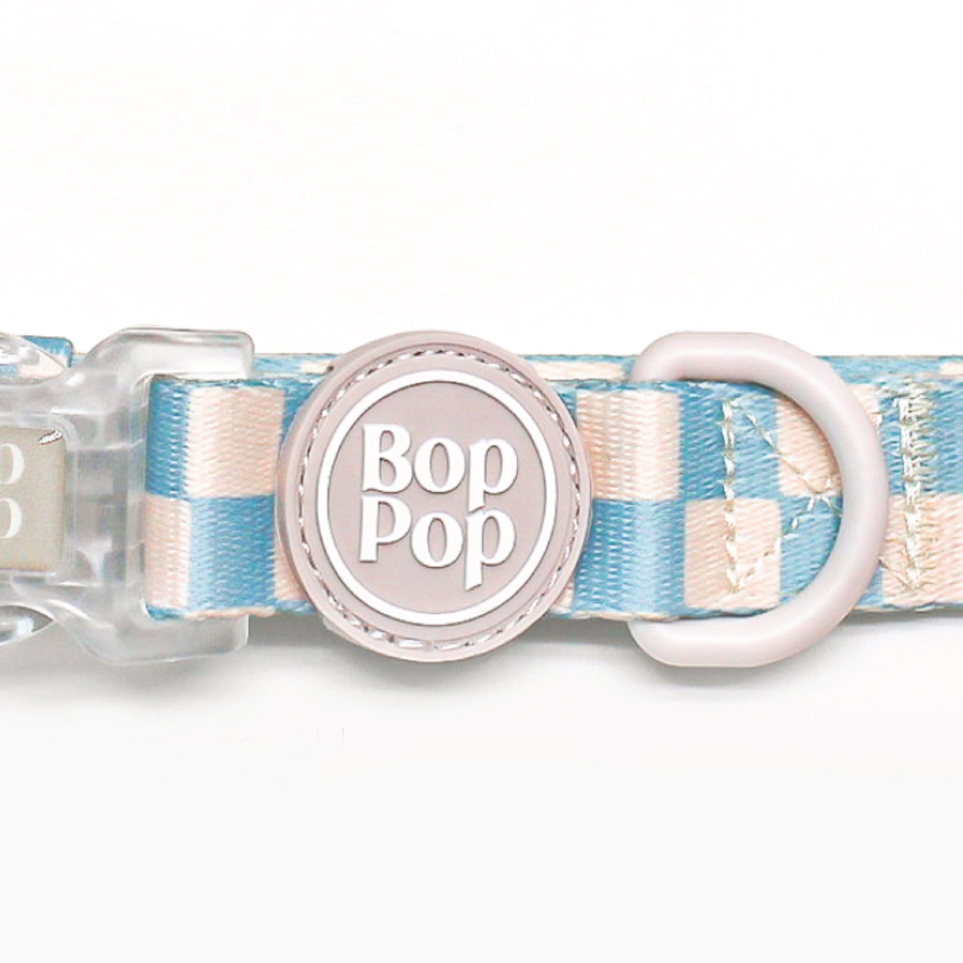 A close-up of a Bop Pop brand dog collar featuring a pastel blue and cream checkered pattern, a light pink plastic D-ring, and a central round pink tag with the brand name 'Bop Pop' in white.