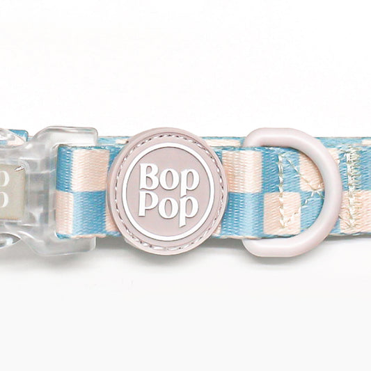A close-up of a Bop Pop brand dog collar featuring a pastel blue and cream checkered pattern, a light pink plastic D-ring, and a central round pink tag with the brand name 'Bop Pop' in white.