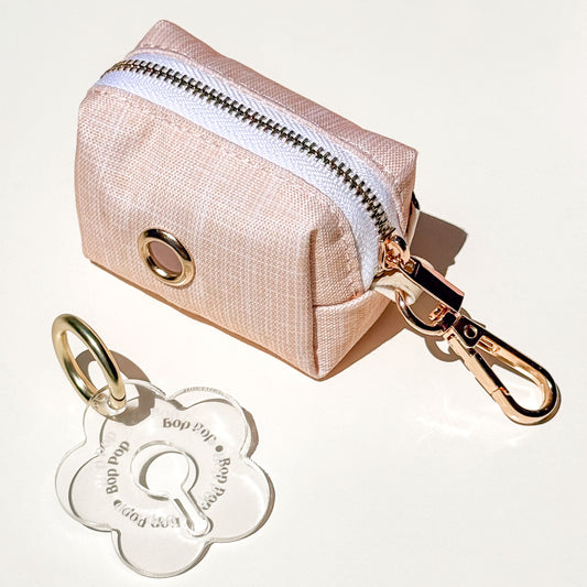 A small boho beige waterproof fabric zipper pouch with a gold-tone metal grommet and attached gold-tone clasp, shown on a white background with a clear flower-shaped poop loop engraved with 'Bop Pop' for dog poop bag dispensing.