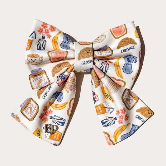 Cotton fabric pet bow featuring a whimsical breakfast food print with croissants, bananas, coffee cups, and peanut butter jars on a cream background, with a small metallic 'BP' monogram charm on left side of sailor bow tail.