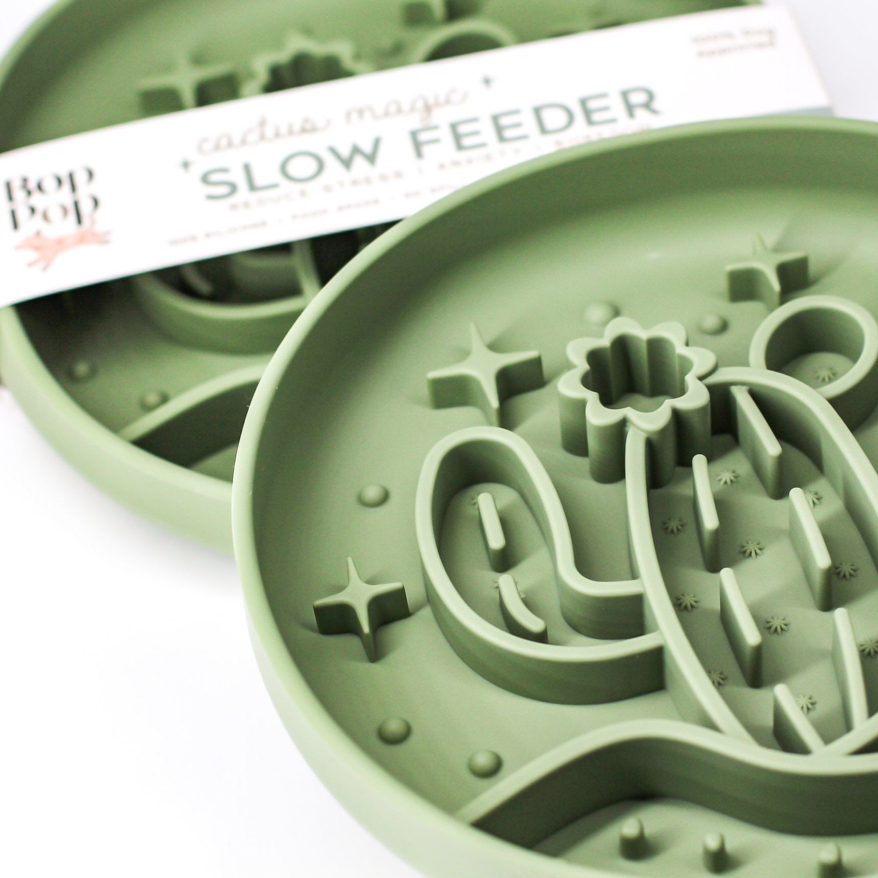 Cactus Magic Slow Feeder staggered on white background with upper left feeder displaying a blurred view of brand packaging labeled 'Cactus Magic SLOW FEEDER' and lower right feeder displaying partially cropped view of 3 dimensional pattern of a cactus with blooming flower in the desert hill with stars in sage green color.
