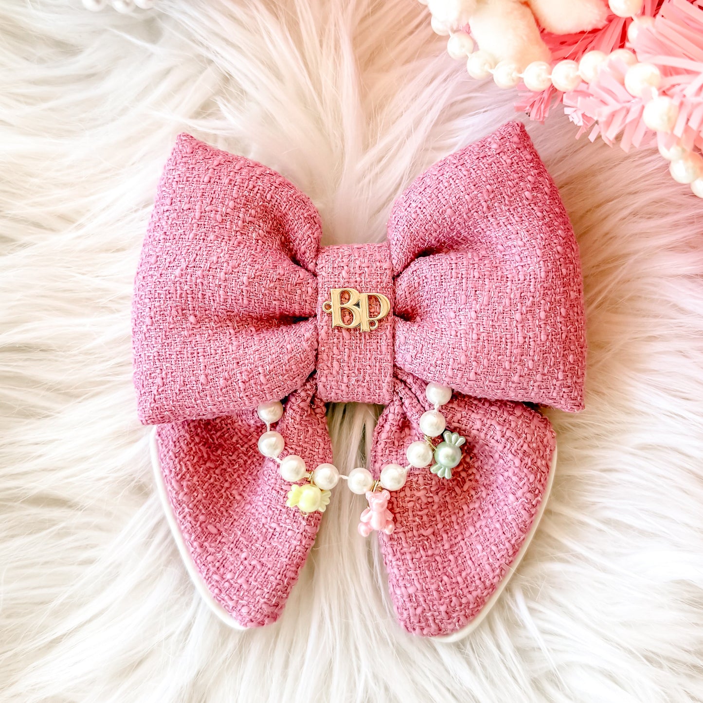 A pink textured fabric dog pet bow tie with a small gold 'BP' gold brand logo sewn on the center, decorated with a short strand of faux pearl beads, displayed on a white faux fur surface with pink decorative props. Miniature charms are teddy bear and pastel candies.