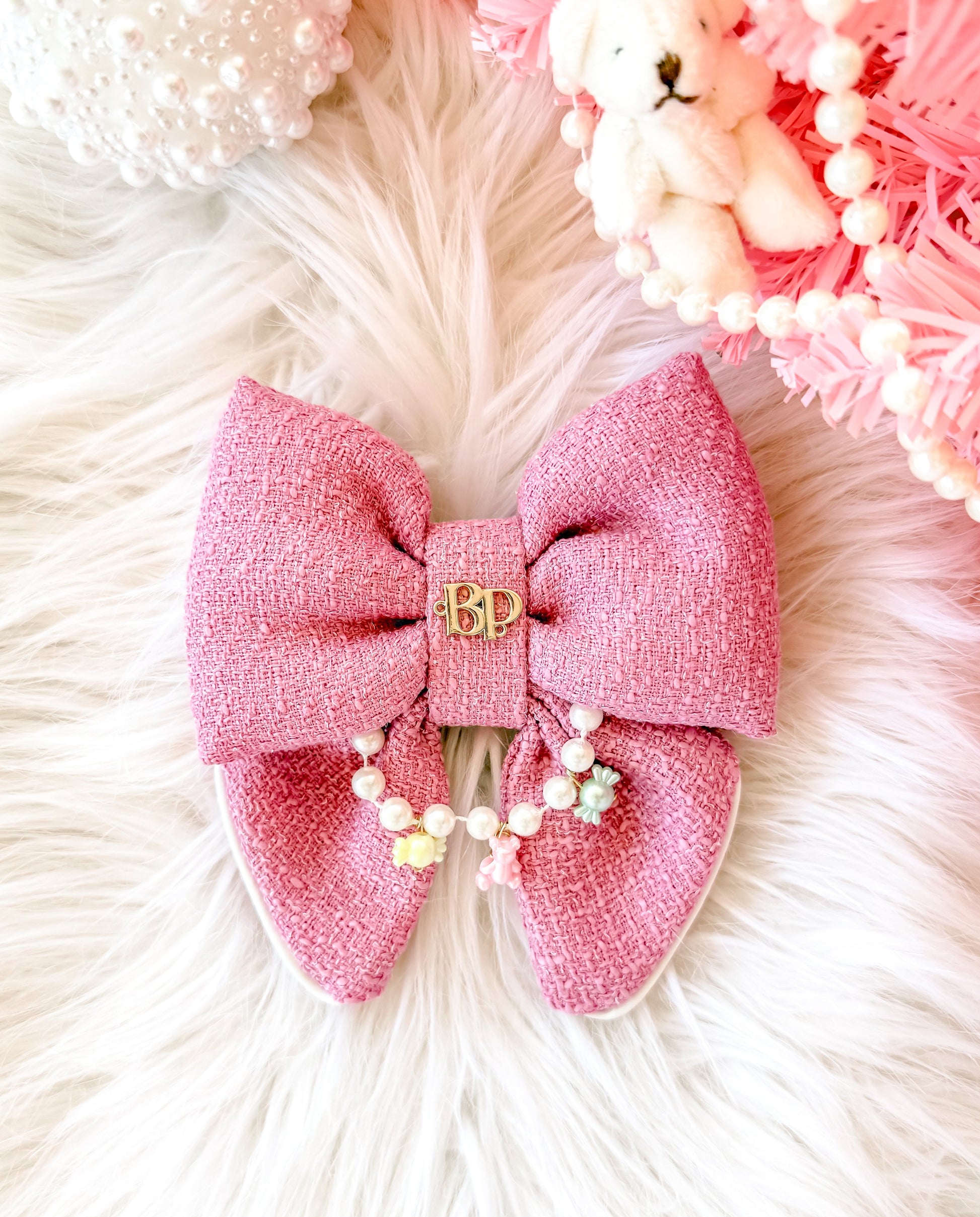 A pink textured fabric dog pet bow tie with a small gold 'BP' gold brand logo sewn on the center, decorated with a short strand of faux pearl beads, displayed on a white faux fur surface with pink decorative props. Miniature charms are teddy bear and pastel candies.