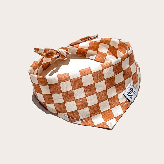 A checkered rust orange and off-white bandana-style pet bandana with a side tag reading 'Bop Pop', photographed on a light beige background.