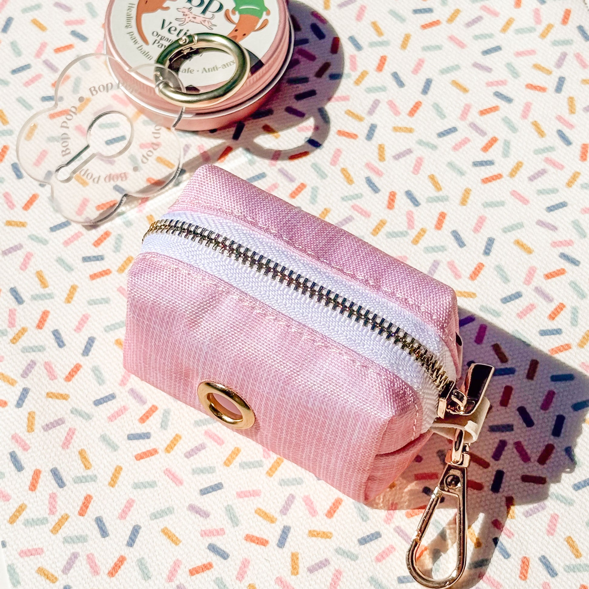 A small pink zippered pouch with a gold-tone snap button and metal clip, photographed on a white surface printed with multicolored confetti. A round pink tin and clear plastic bag are partially visible in the background.