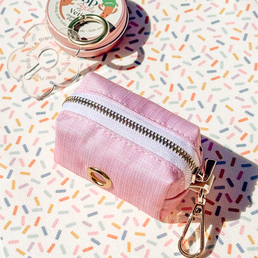 A small pink zippered pouch with a gold-tone snap button and metal clip, photographed on a white surface printed with multicolored confetti. A round pink tin and clear plastic bag are partially visible in the background.