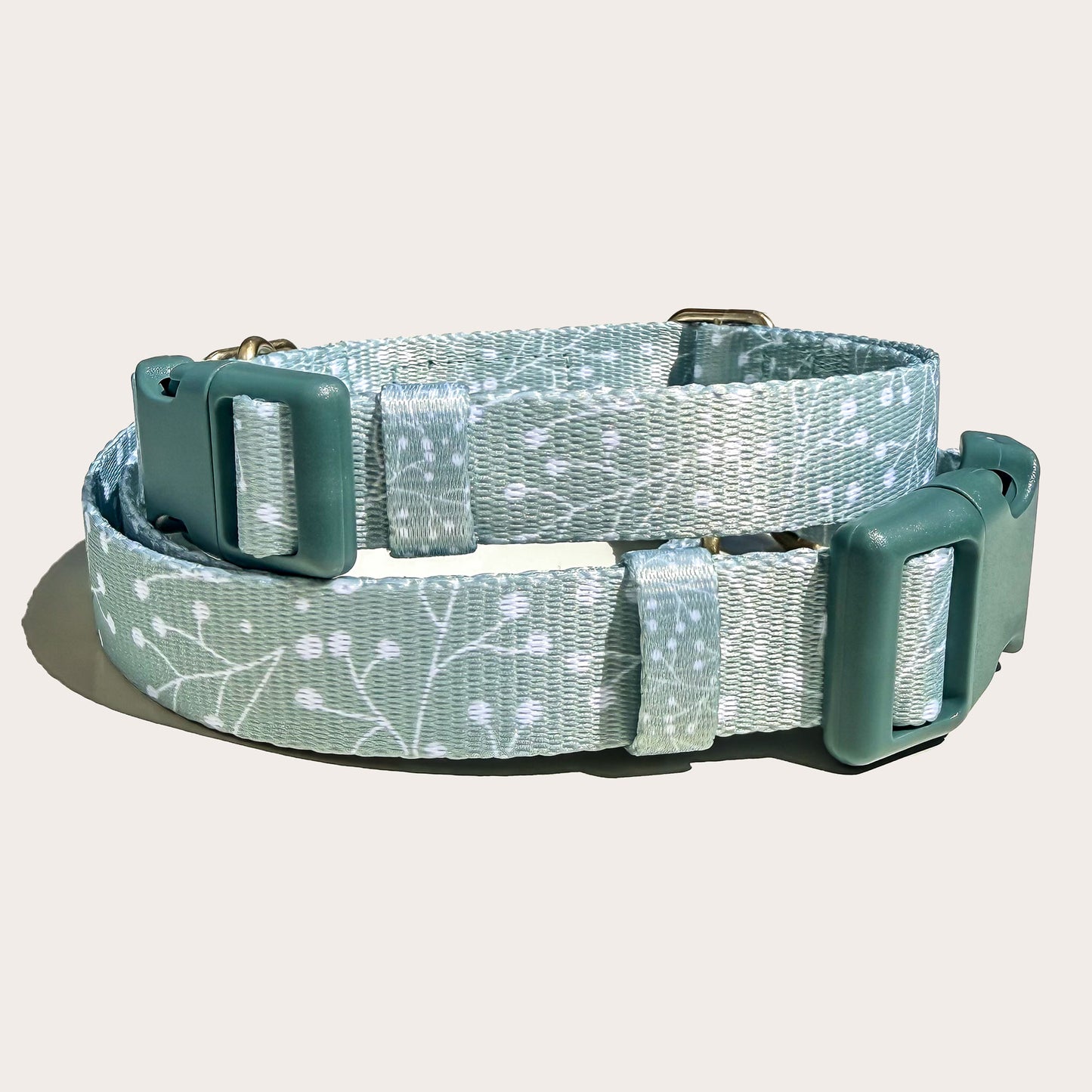 Two stacked dog collars, top one smaller with a sage green and white floral juniper plant branch pattern, featuring dark sage green plastic buckles and metal hardware, shown on a light off-white background.