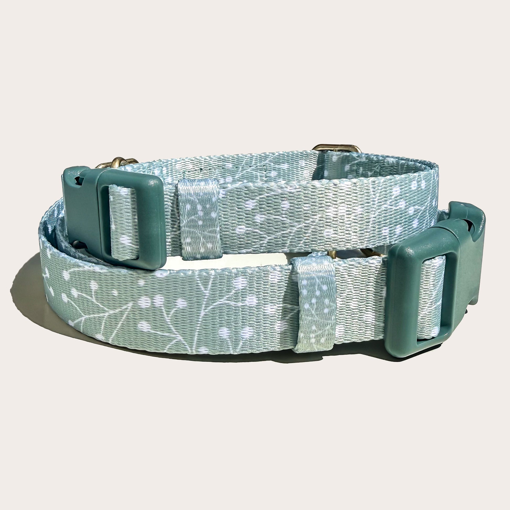 Two stacked dog collars, top one smaller with a sage green and white floral juniper plant branch pattern, featuring dark sage green plastic buckles and metal hardware, shown on a light off-white background.