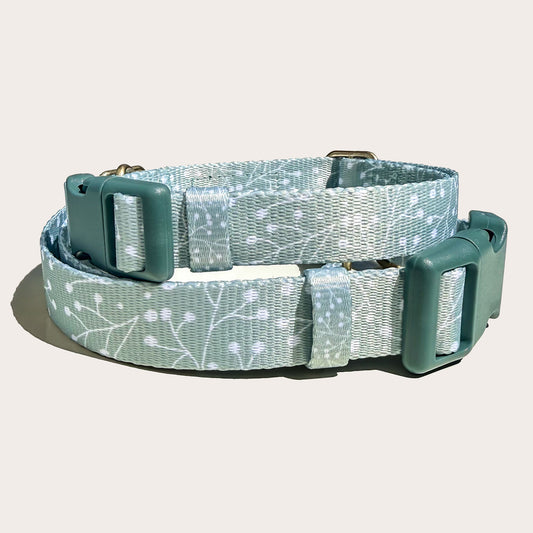 Two stacked dog collars, top one smaller with a sage green and white floral juniper plant branch pattern, featuring dark sage green plastic buckles and metal hardware, shown on a light off-white background.