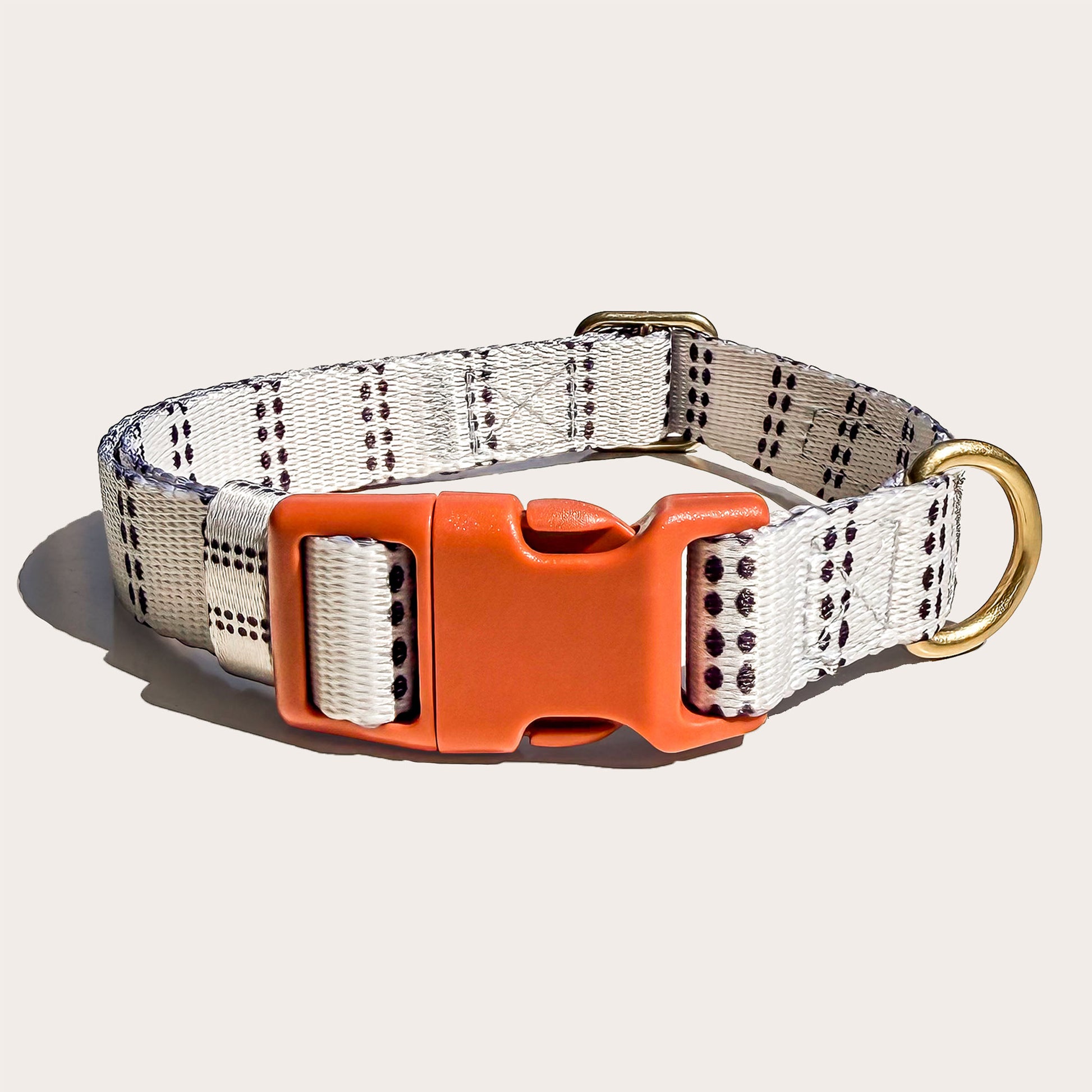 A dog collar with a white woven strap featuring black dotted grid patterns, an orange plastic quick-release buckle, and a gold-tone metal ring, shown on a light beige background.