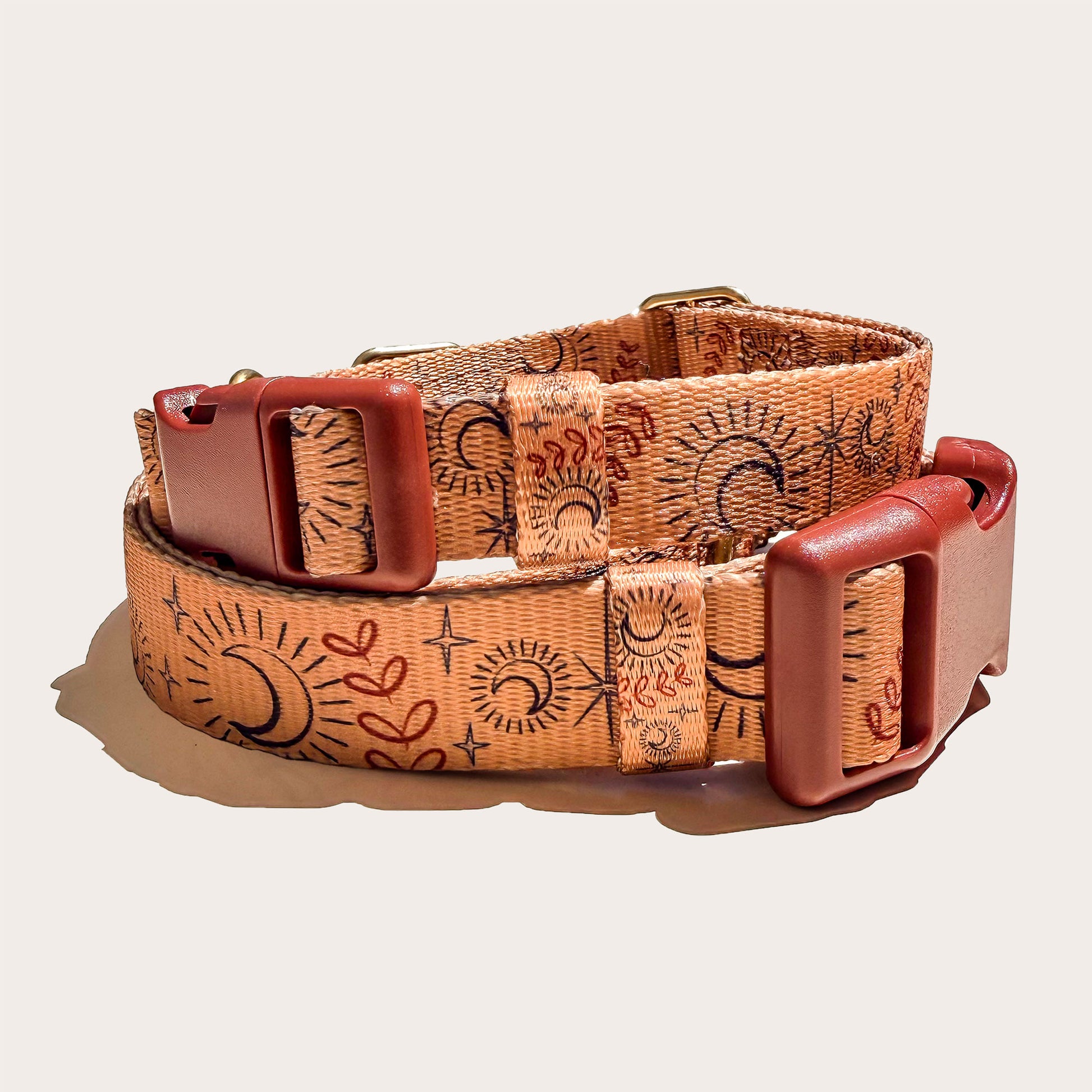 A celestial stlyed boho dog collar with a warm tan base, printed with black and reddish-brown sun and moon motifs, and finished with reddish-brown leather buckles, photographed on a light beige background.