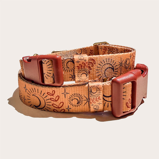 A celestial stlyed boho dog collar with a warm tan base, printed with black and reddish-brown sun and moon motifs, and finished with reddish-brown leather buckles, photographed on a light beige background.