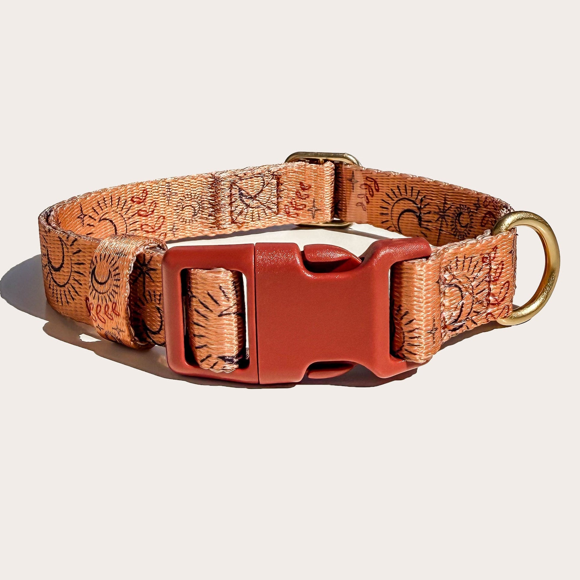 A dog collar with a warm orange and brown patterned webbing, featuring a prominent red plastic buckle and a brass ring, shown on a light beige background.