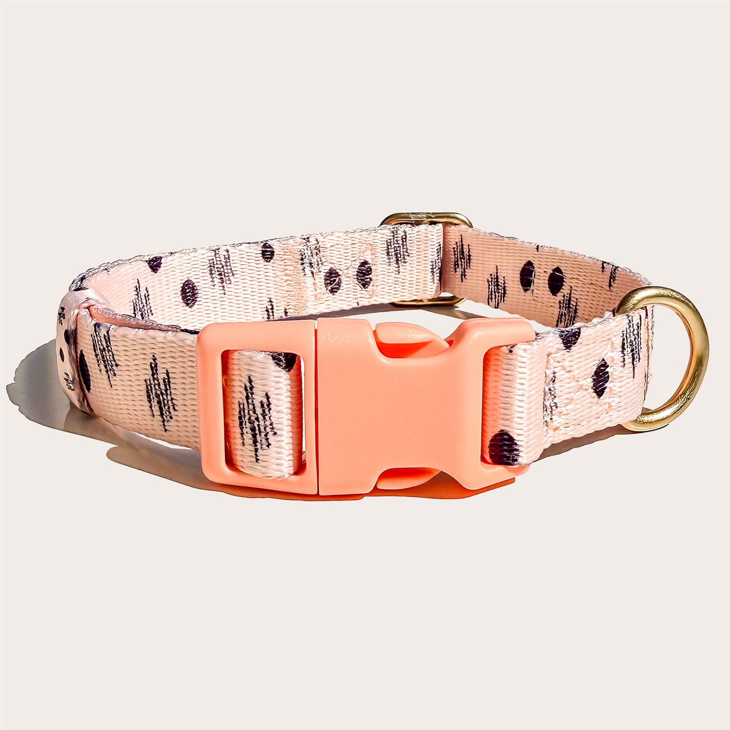 A pink peach dog collar with a black ikat abstract pattern, featuring a bright coral orange plastic buckle and a gold-tone metal ring, shown on a light beige background.
