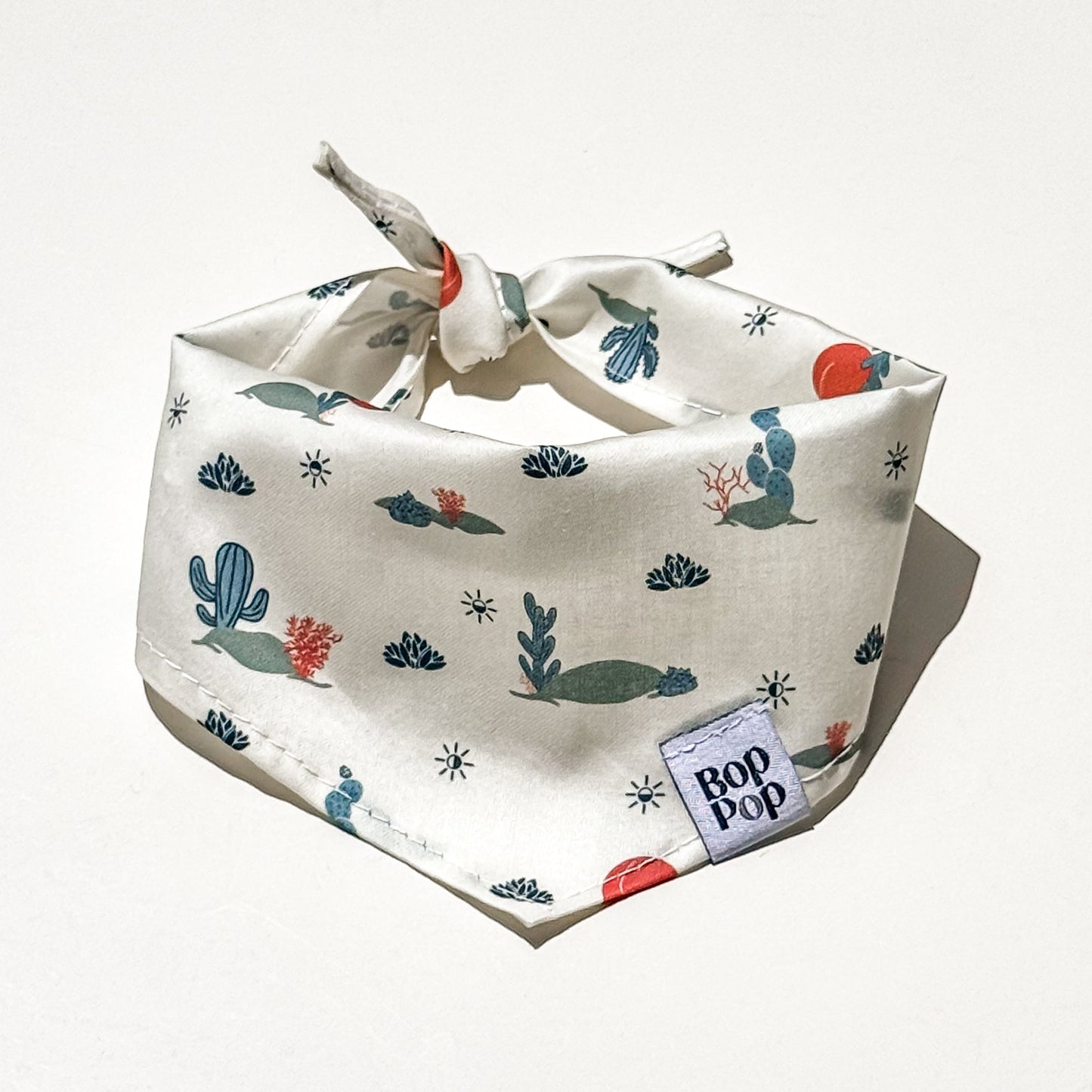 A cream-colored dog bandana with a tie-loop design, featuring a whimsical desert print of cacti, succulents, and small blue birds. A small woven label on the corner reads 'BOP POP'.