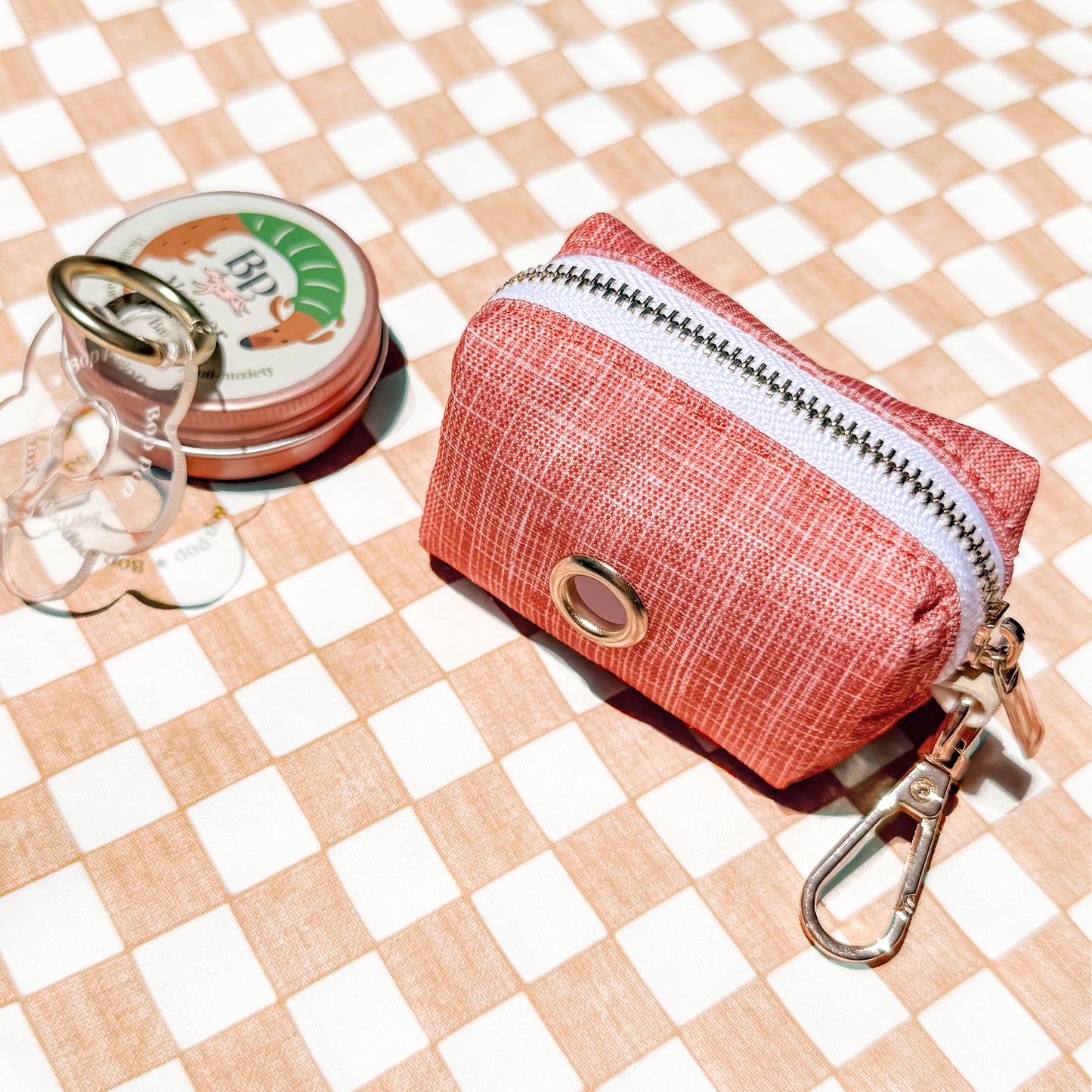 A small zippered pouch in a coral orange textured fabric with a white zipper, gold-tone metal grommet, and attached gold keychain clasp, placed on a beige and white checkered surface. A round tin and a clear flower-shaped accessory are partially visible beside it.