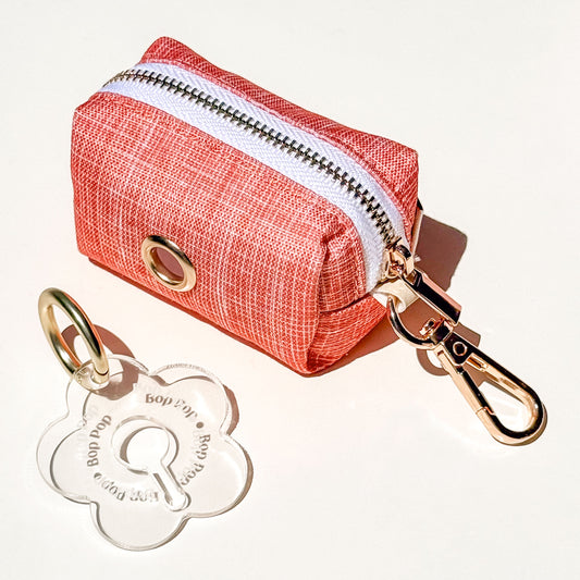 A small zippered pouch in a textured coral orange waterproof fabric with a gold-tone metal grommet and attached gold-tone clasp, shown on a white background with a clear flower-shaped poop loop engraved with 'Bop Pop' for dog poop bag dispensing.
