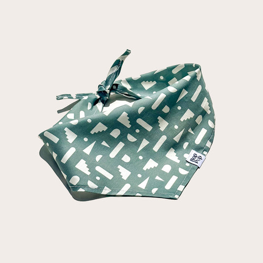 A sage green pet bandana with a white geometric pattern, tied in a bow and featuring a small white brand tag that reads 'BOP POP'  shown on a light beige background.