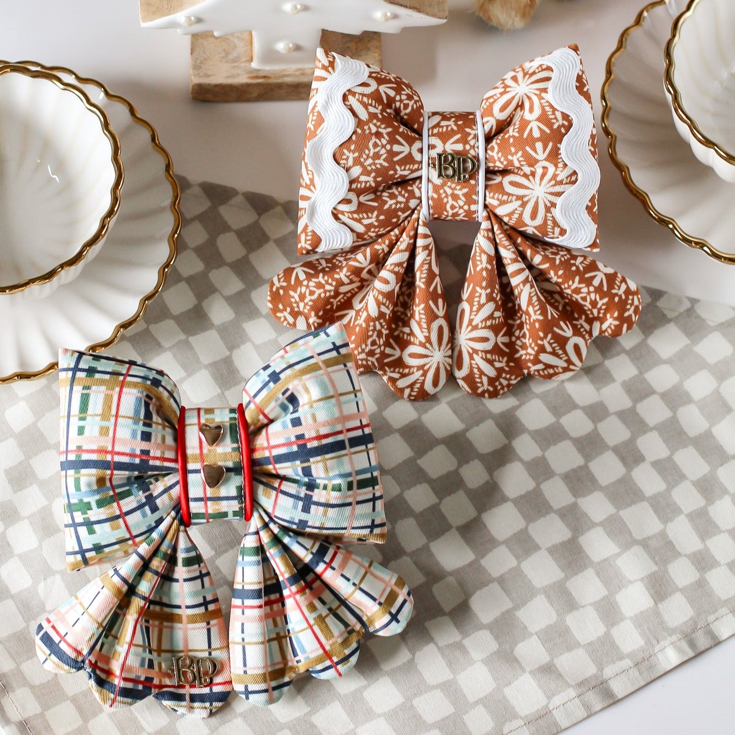 Two decorative bows on a checkered tablecloth with a small wooden tree. Gingerbread and Plaid cotton dog sailor bows with gold metal hearts and ric rac. Scallop details for tails.