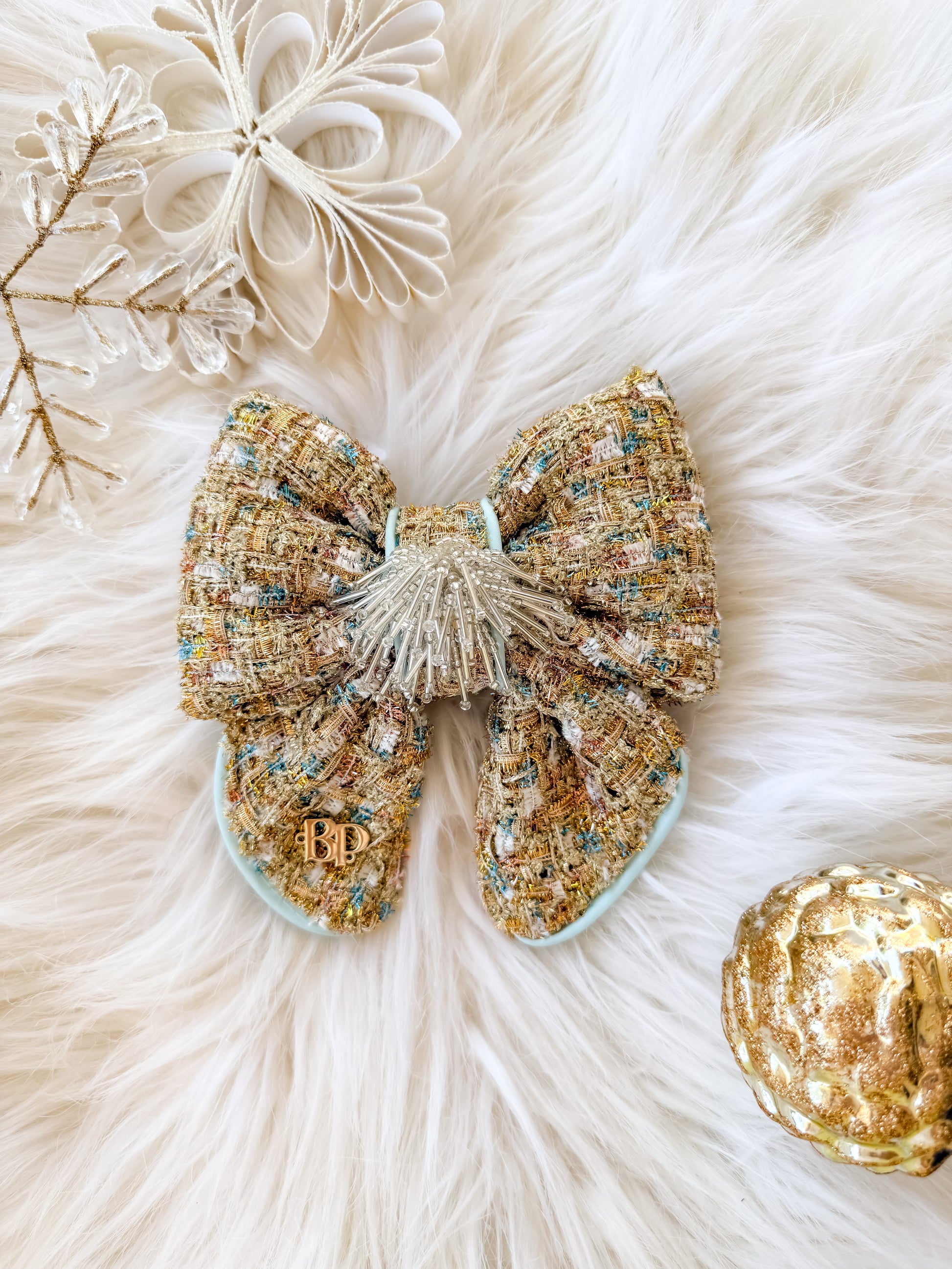 A large decorative dog bow tie with tails made of textured tweed fabric in gold, and aqua blue accent, with a decorative sequin tassel center and a small gold brand logo 'BP' laid on a white faux fur background with holiday decorations.