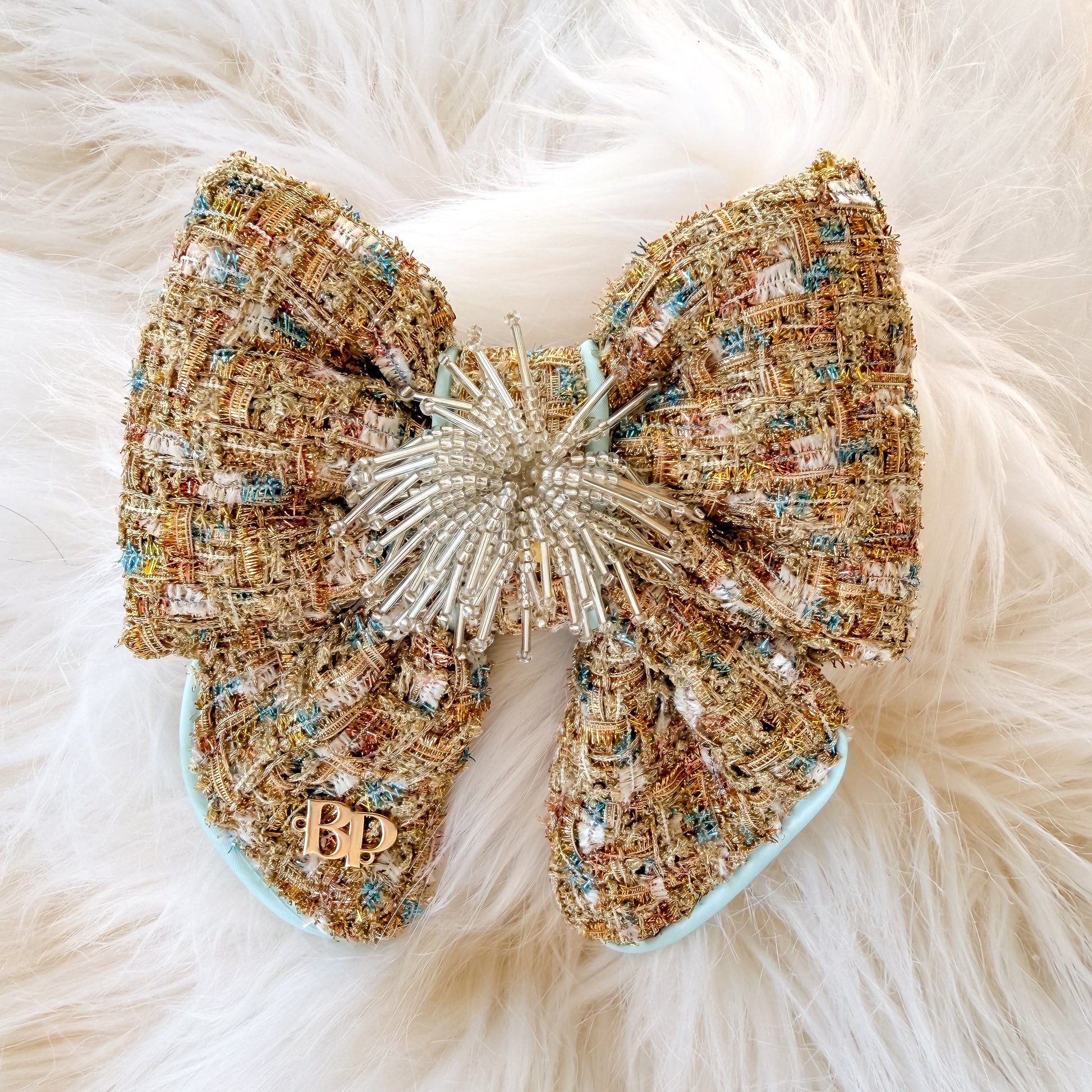 Gold decorative dog bow tie with tails made of textured tweed fabric in beige, gold, and light blue satin trim, with a beaded starburst center and a small gold brand logo 'BP' detail on one loop, placed on a white faux-fur background.