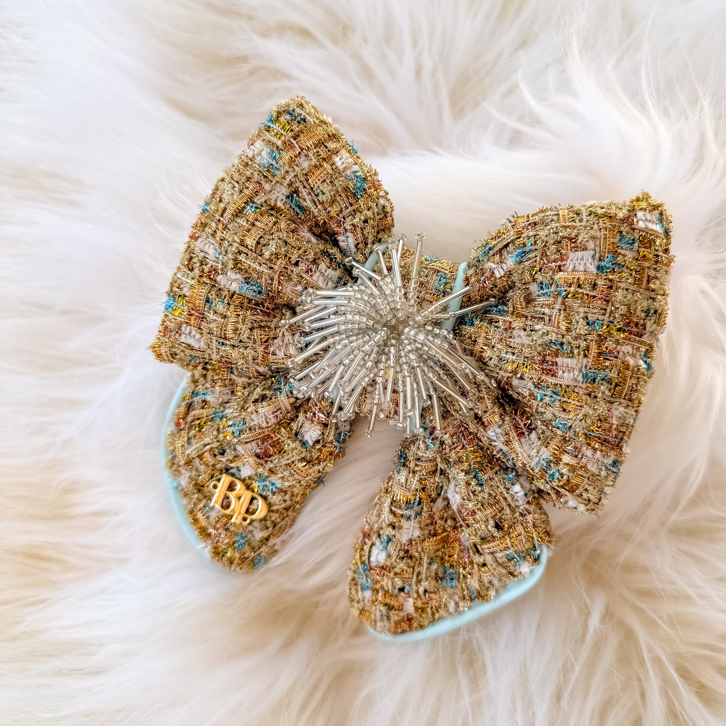 Gold decorative dog bow tie with tails made of textured tweed fabric in beige, gold, and light blue satin trim, with a beaded starburst center and a small gold brand logo 'BP' detail on one loop, placed on a white faux-fur background.