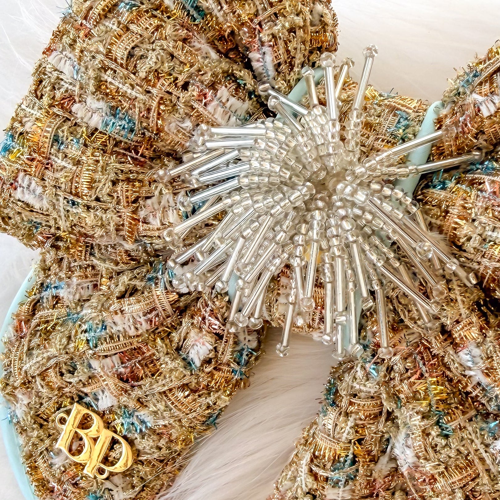 Gold decorative dog bow tie with tails made of textured tweed fabric in beige, gold, and light blue satin trim, with a beaded starburst center and a small gold brand logo 'BP' detail on one loop, placed on a white faux-fur background.
